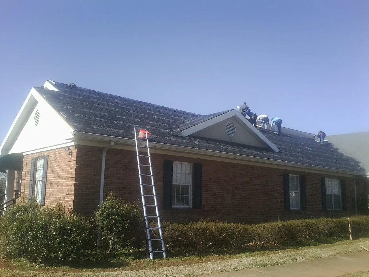 Crew tearing shingles off a brick ranch roof with a ladder set — Metal Roof Repair work by our crew in Herminie
